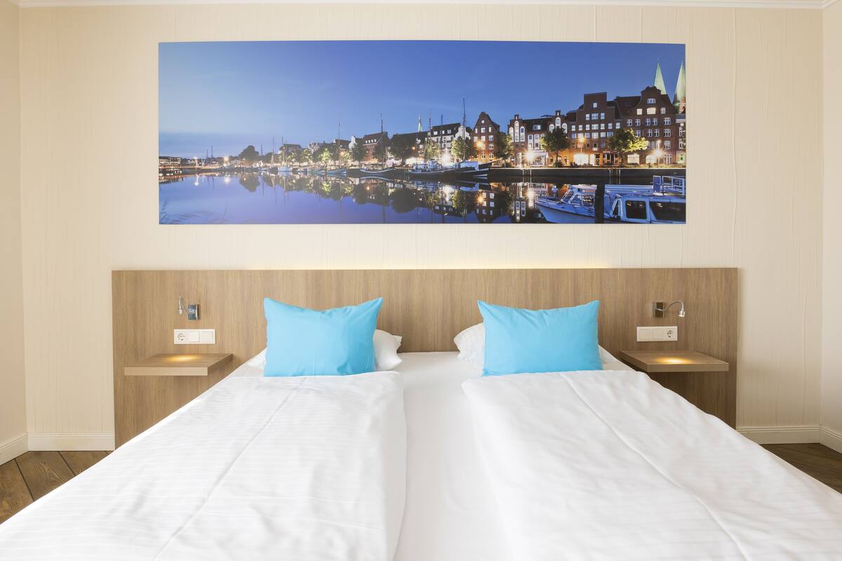 Una cama doble con sábanas blancas y dos almohadas azules, un cabecero de madera y una mesita de noche con lámpara.