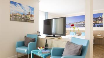 Ein modernes Hotelzimmer mit einem großen Flachbildfernseher, zwei blauen Sesseln, einem kleinen Tisch mit Flasche und Gläsern sowie Blick auf die Stadt.