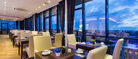 Un restaurante moderno con techos altos, amplias ventanas y vista a la ciudad. Las mesas están ordenadas con sillas, y el suelo es de madera.