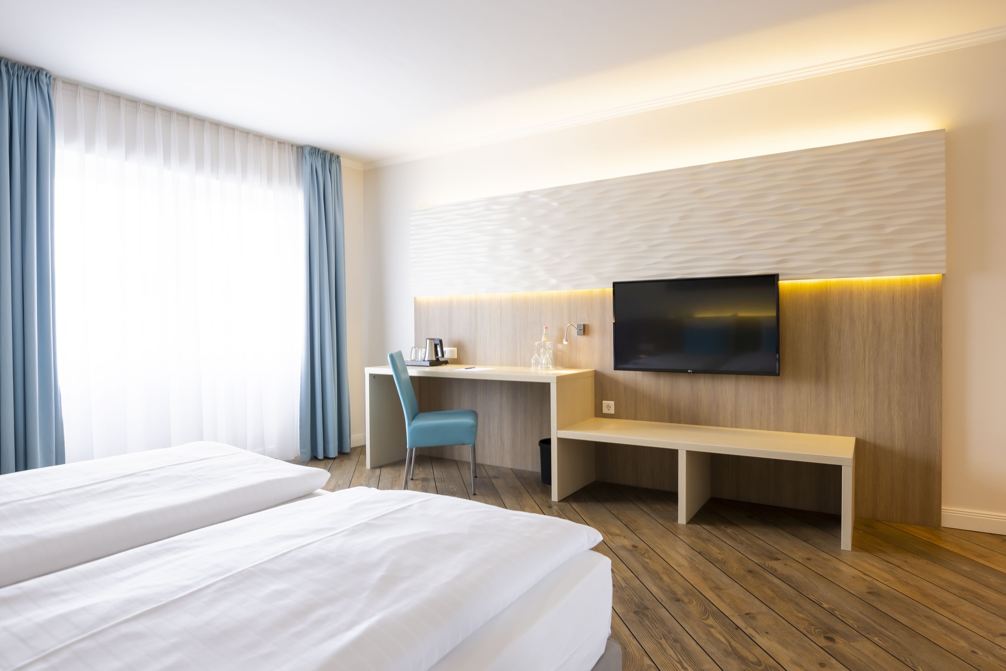 A hotel room with two beds, a desk, a chair, a TV, and a bench.