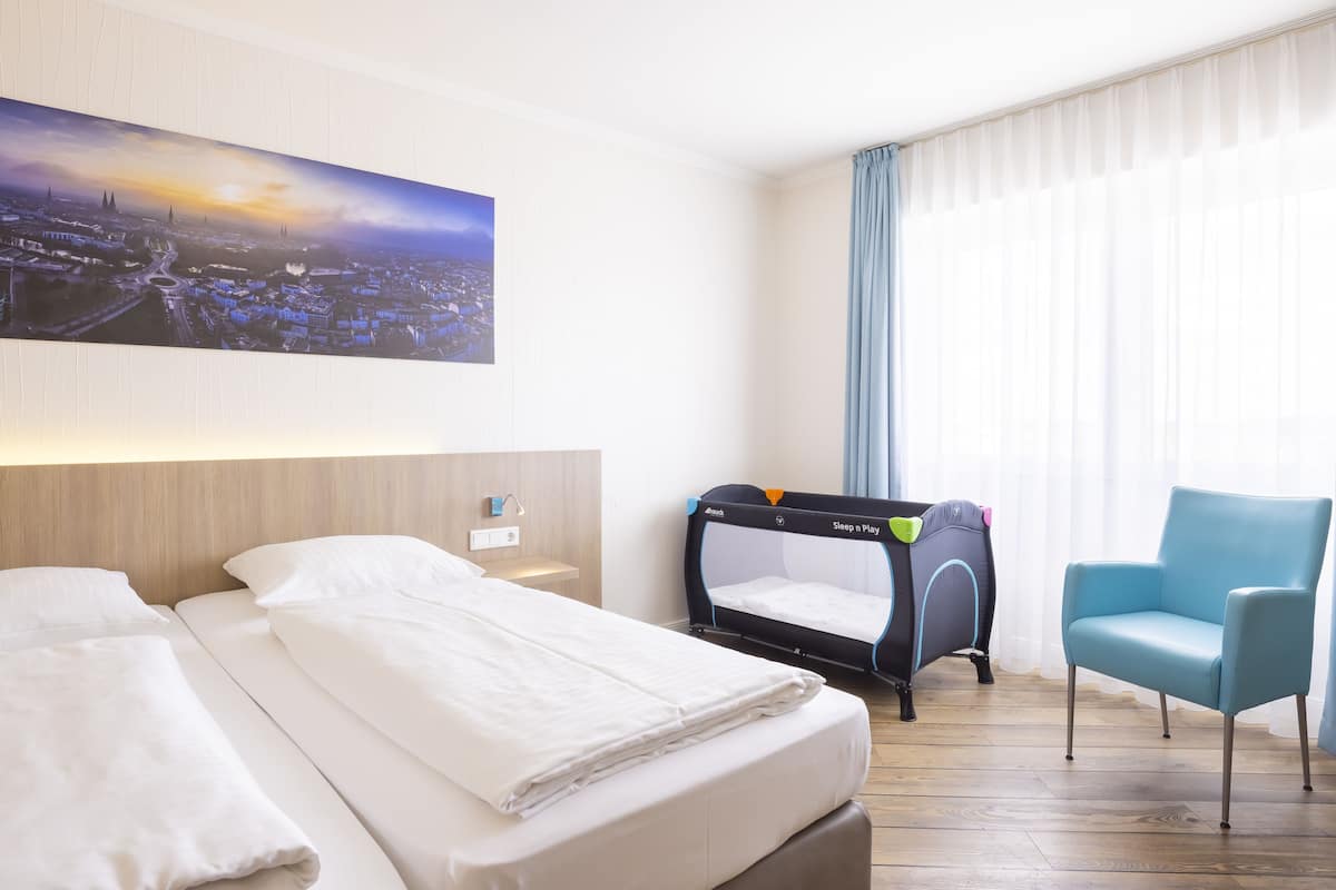 Habitación de hotel con cama, cuna, silla y una pintura de paisaje urbano en la pared.