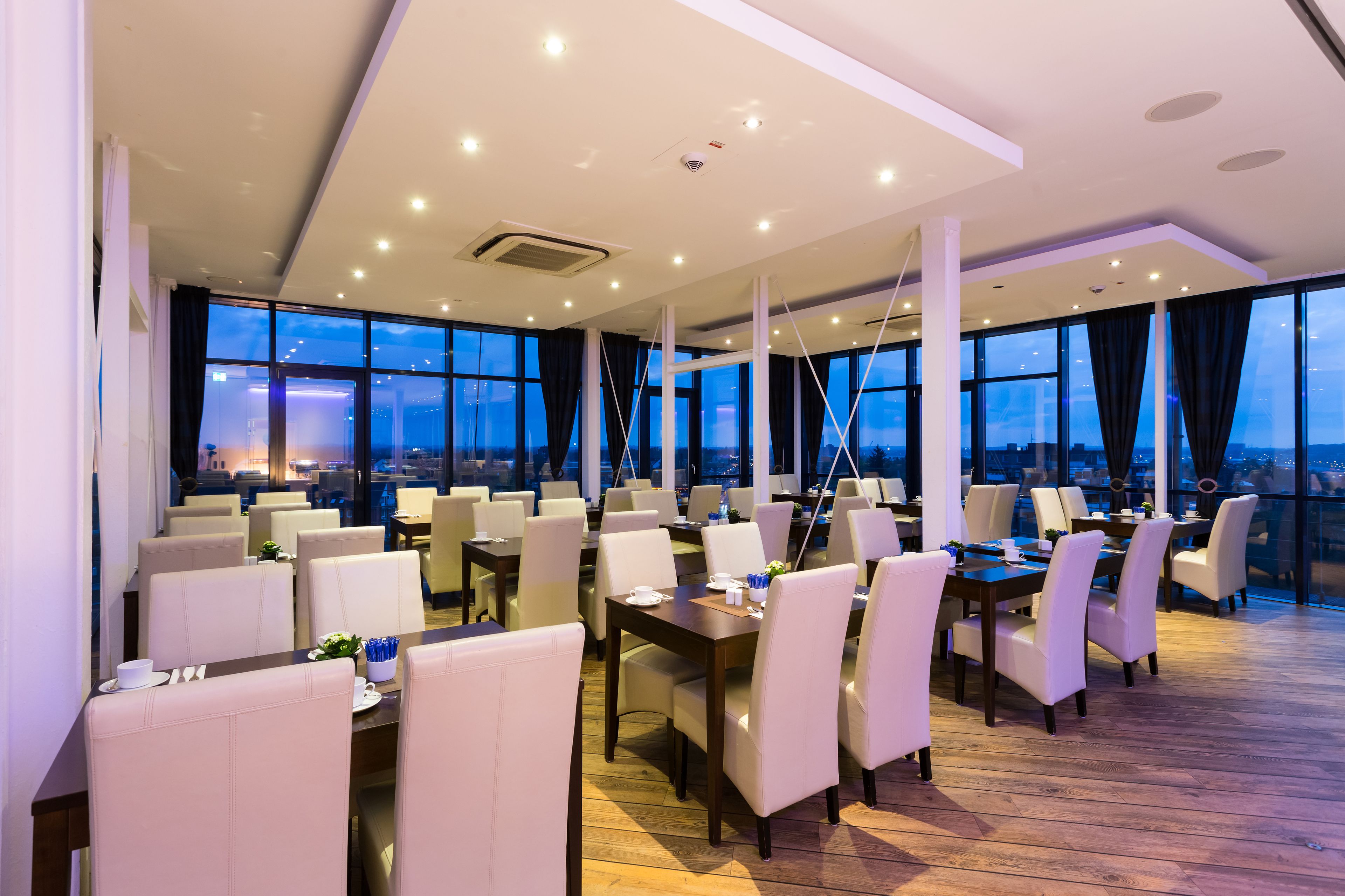A modern dining area with multiple tables and chairs, large windows offering a city view, and a wooden floor.