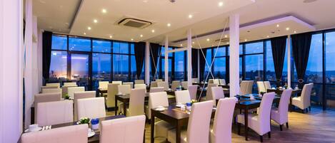 A modern dining area with multiple tables and chairs, large windows offering a city view, and a wooden floor.