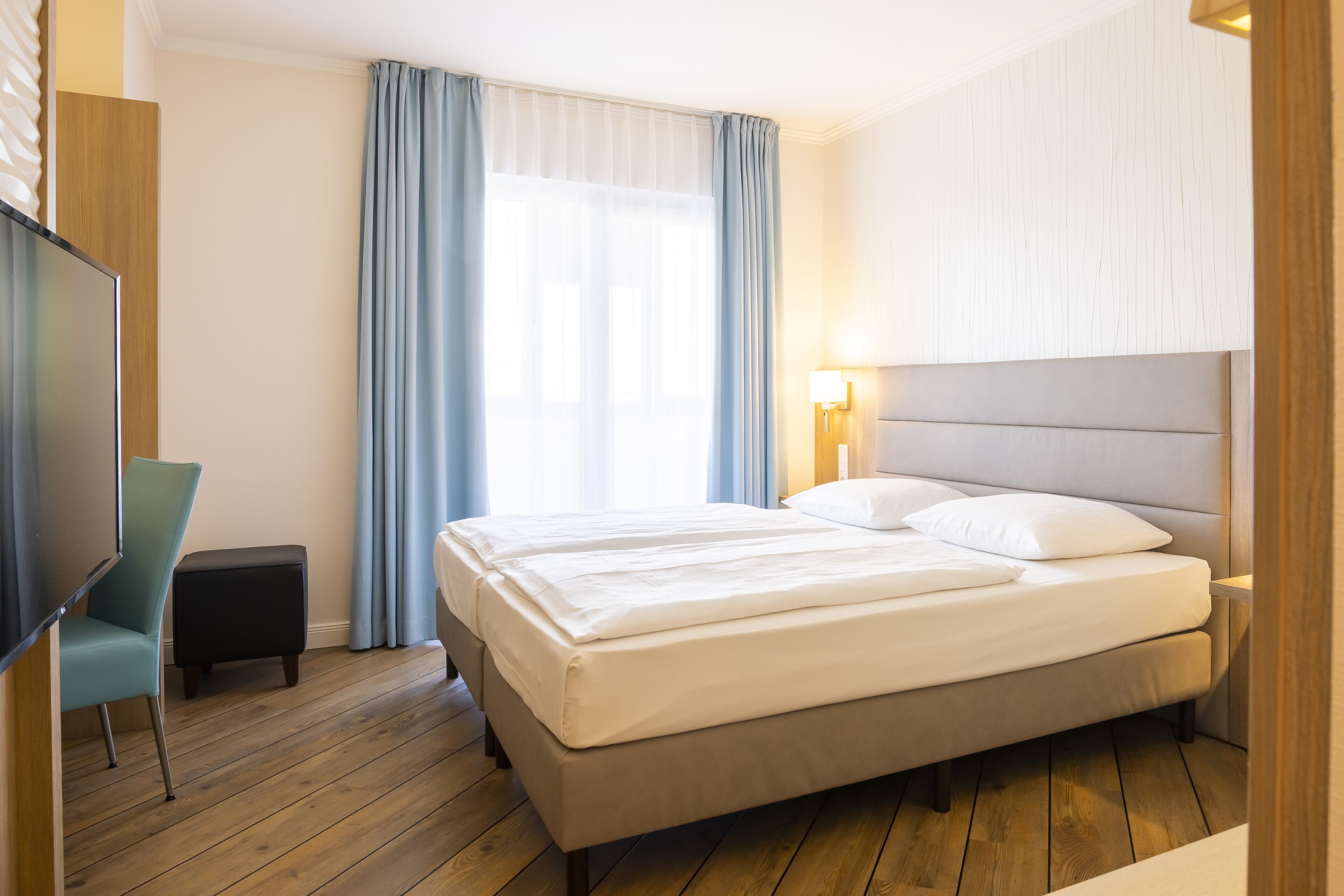 A hotel room with a large bed, a TV, a small table, and a window with curtains.