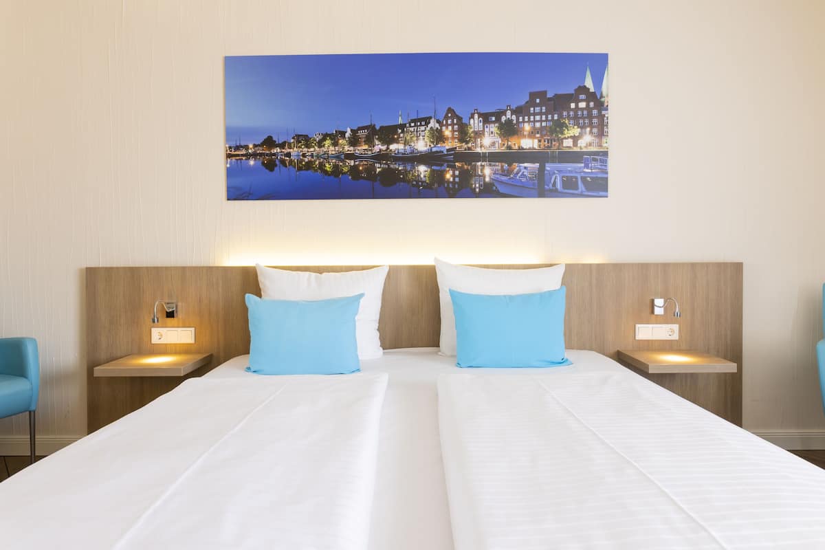 Habitación de hotel con dos camas, una mesita de noche con lámpara y un cuadro en la pared de un paisaje urbano.