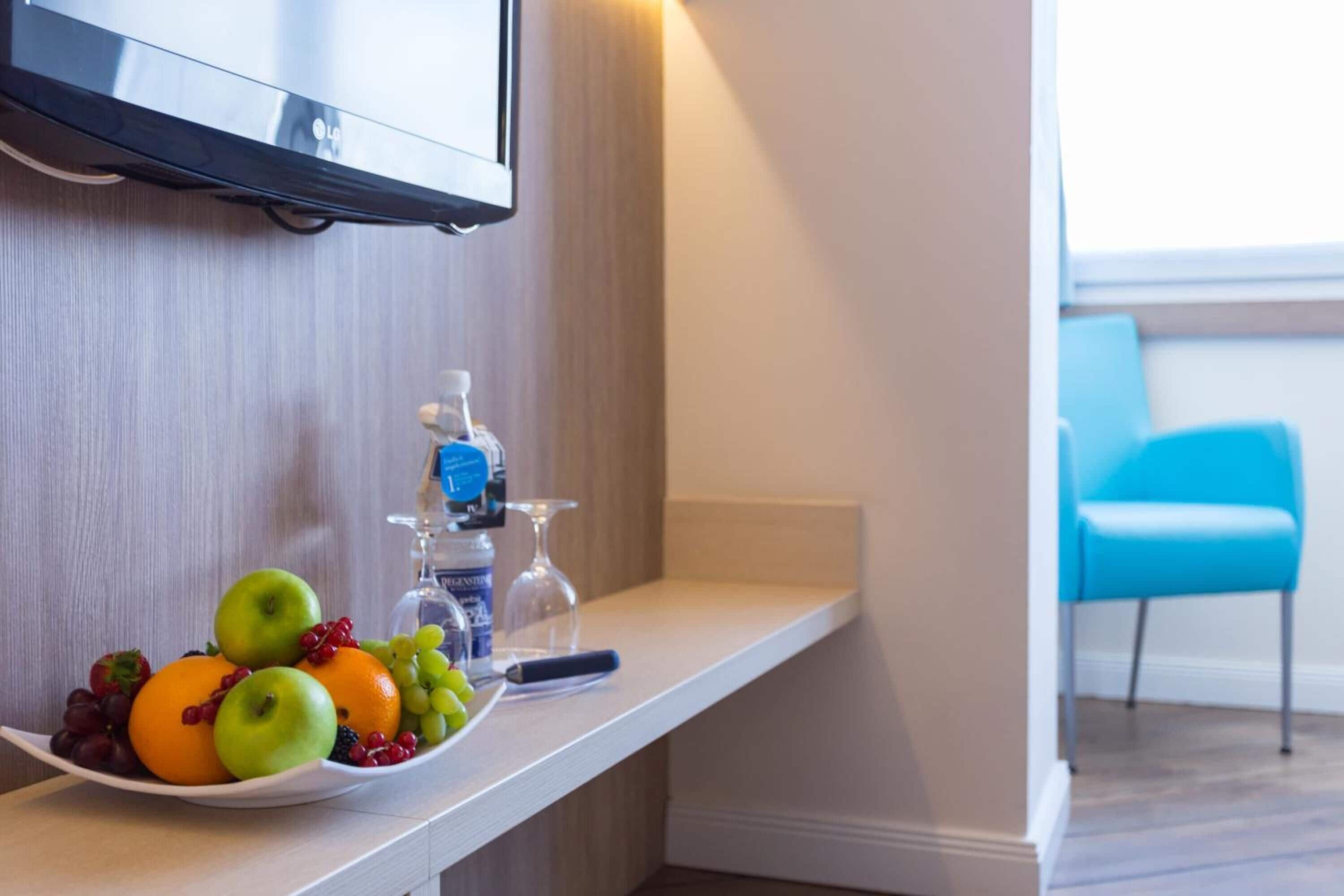 Habitación de hotel con una consola blanca que exhibe frutas y una botella de agua, un televisor montado en la pared y una silla azul en la esquina.