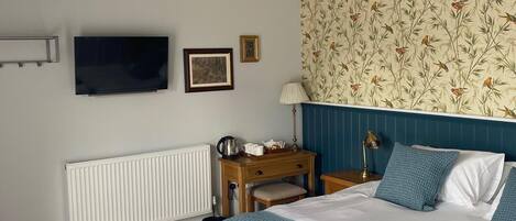 A bedroom with a large bed, a wooden nightstand, a desk, a radiator, a TV, and a patterned wallpaper.