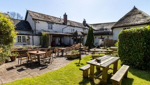 An outdoor patio area with wooden tables and chairs, surrounded by a white building with a thatched roof. - The New Inn (Crediton)