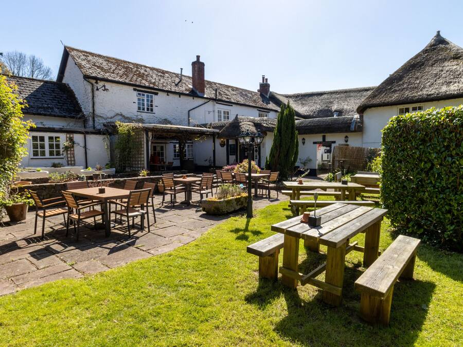 An outdoor patio area with wooden tables and chairs, surrounded by a white building with a thatched roof.