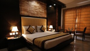 A spacious bedroom with a large bed, two bedside tables with lamps, a wooden headboard, and a view of a cityscape through a large window. - Hotel Delhi Pride (New Delhi)