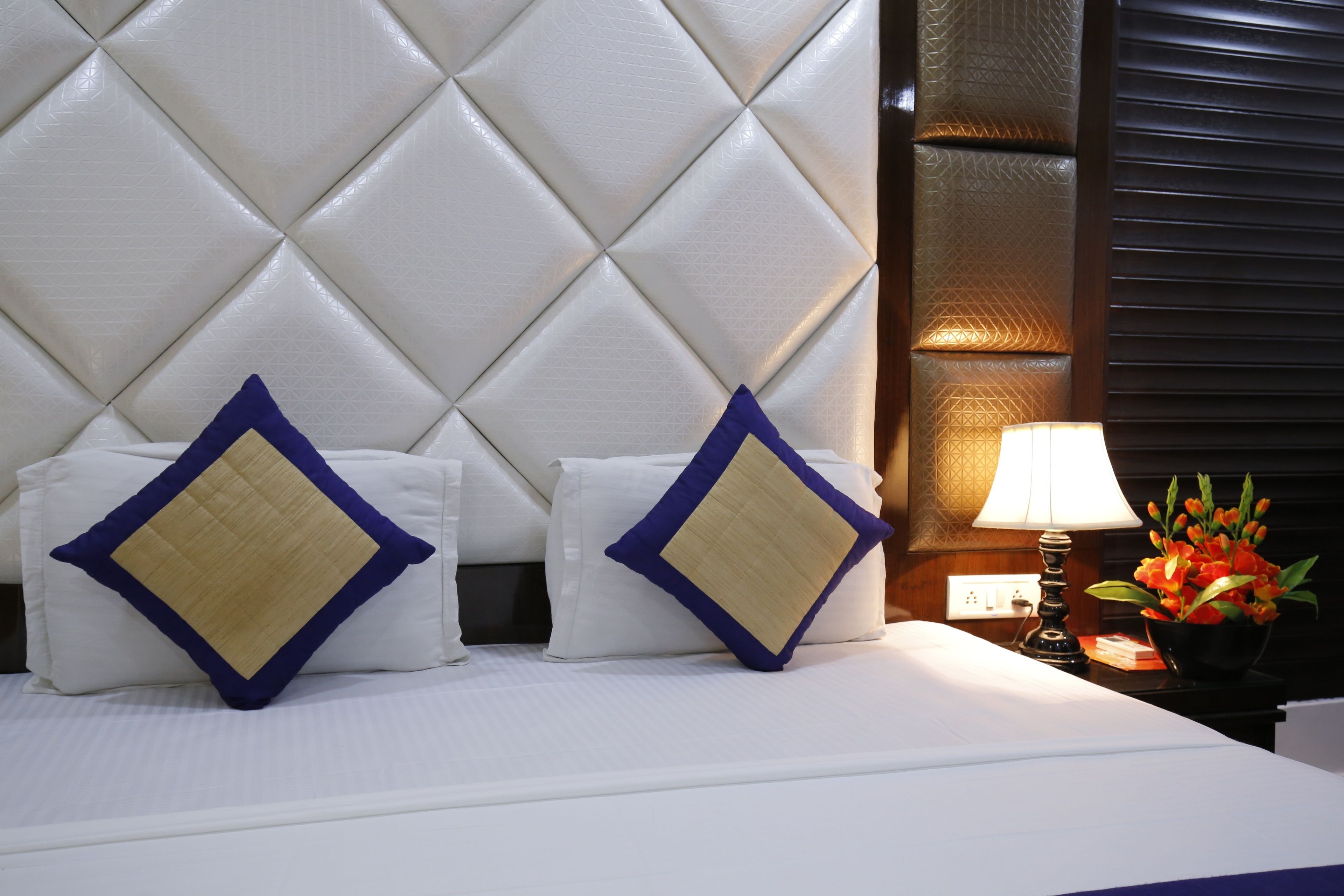 A hotel room with a tufted headboard, two pillows with a blue and beige design, a bedside lamp, and a flower arrangement.