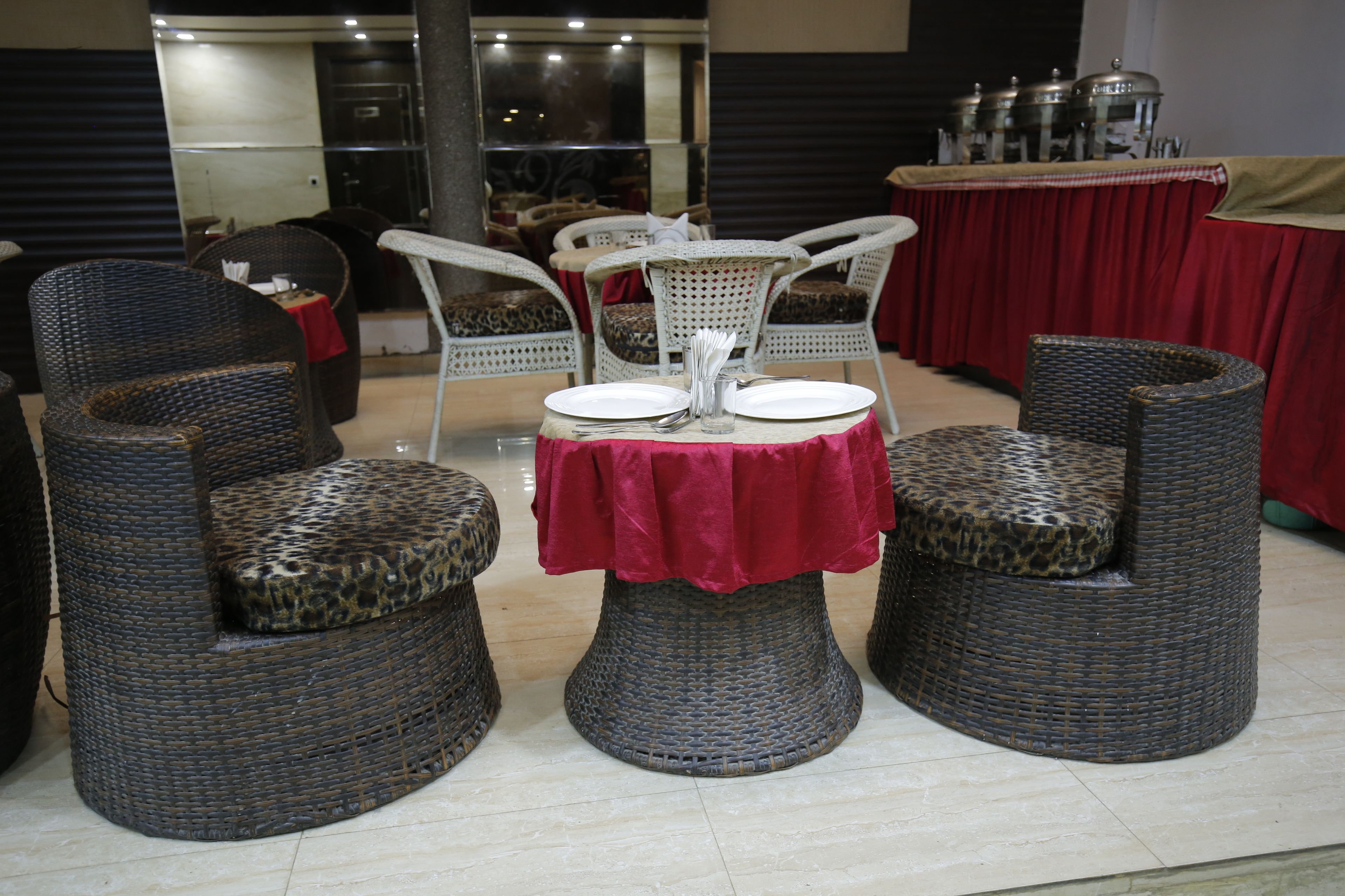Un coin repas avec des chaises en osier et une table dressée pour deux personnes, agrémentée d’une nappe rouge.