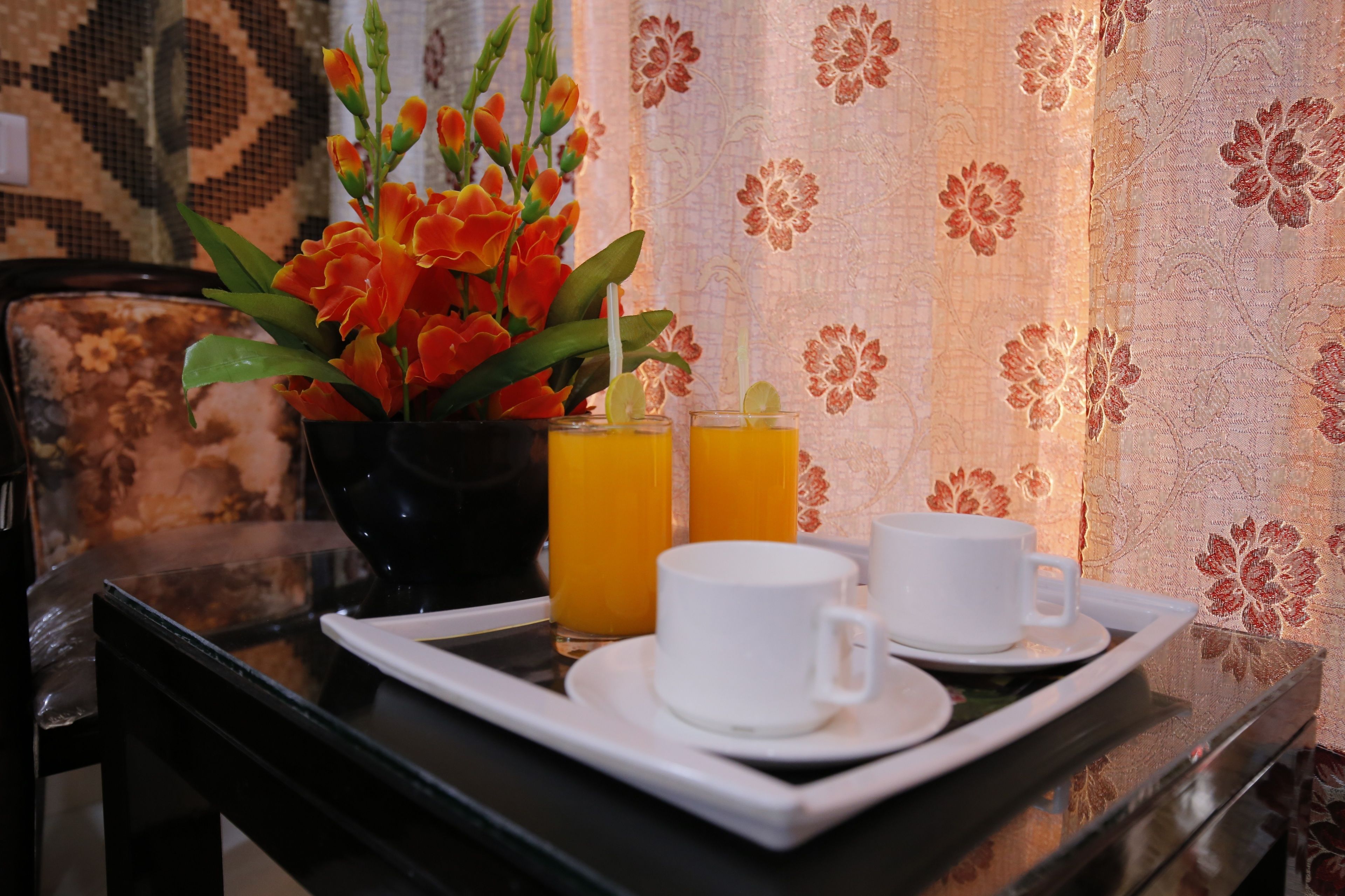A tray with two white cups and a glass of orange juice, a vase with orange flowers, and a patterned wall in the background.