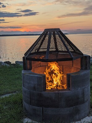 A circular fire pit with a metal lattice cover, surrounded by grass and a body of water in the background. - Motel Rimouski (Rimouski)