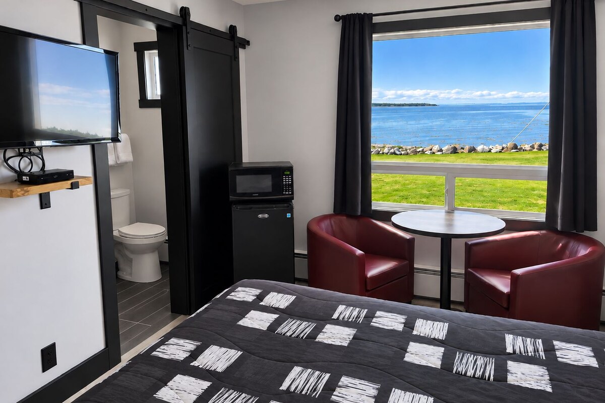 A hotel room with a bed, a TV, a microwave, a small fridge, a toilet, and a window with a view of the sea.