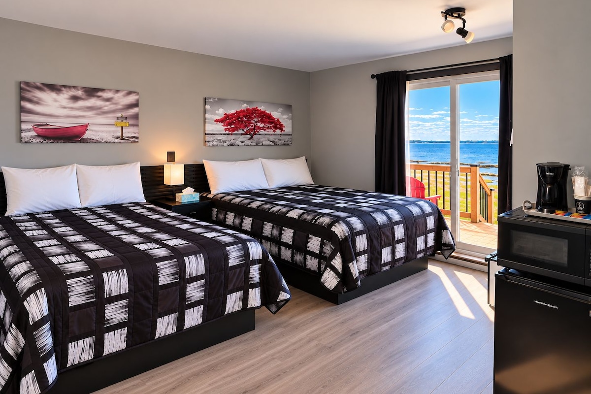 A hotel room with two beds, a nightstand, a lamp, a coffee maker, and a view of the sea through a sliding door.