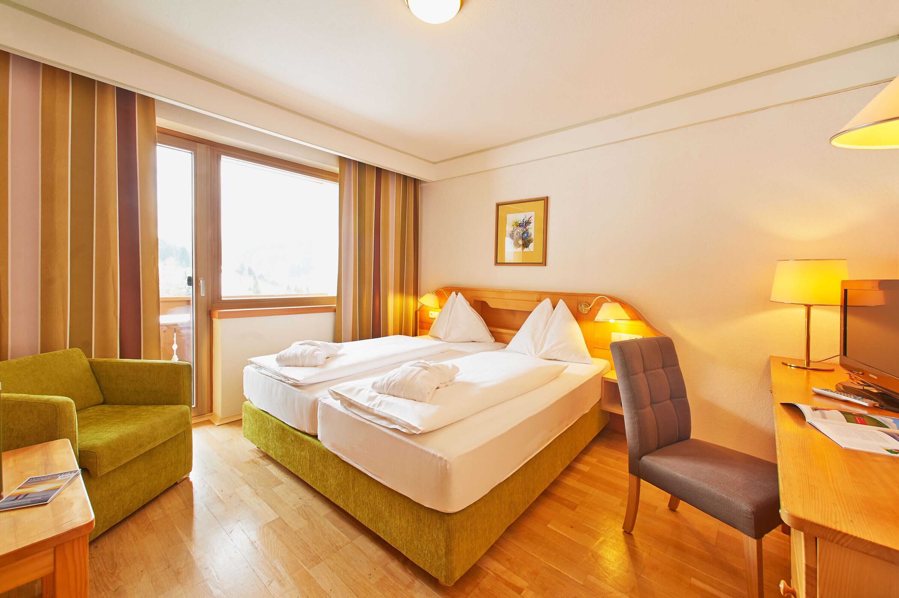 Quarto de hotel com cama, duas mesas de cabeceira, uma poltrona verde, escrivaninha com luminária e janela com cortinas.
