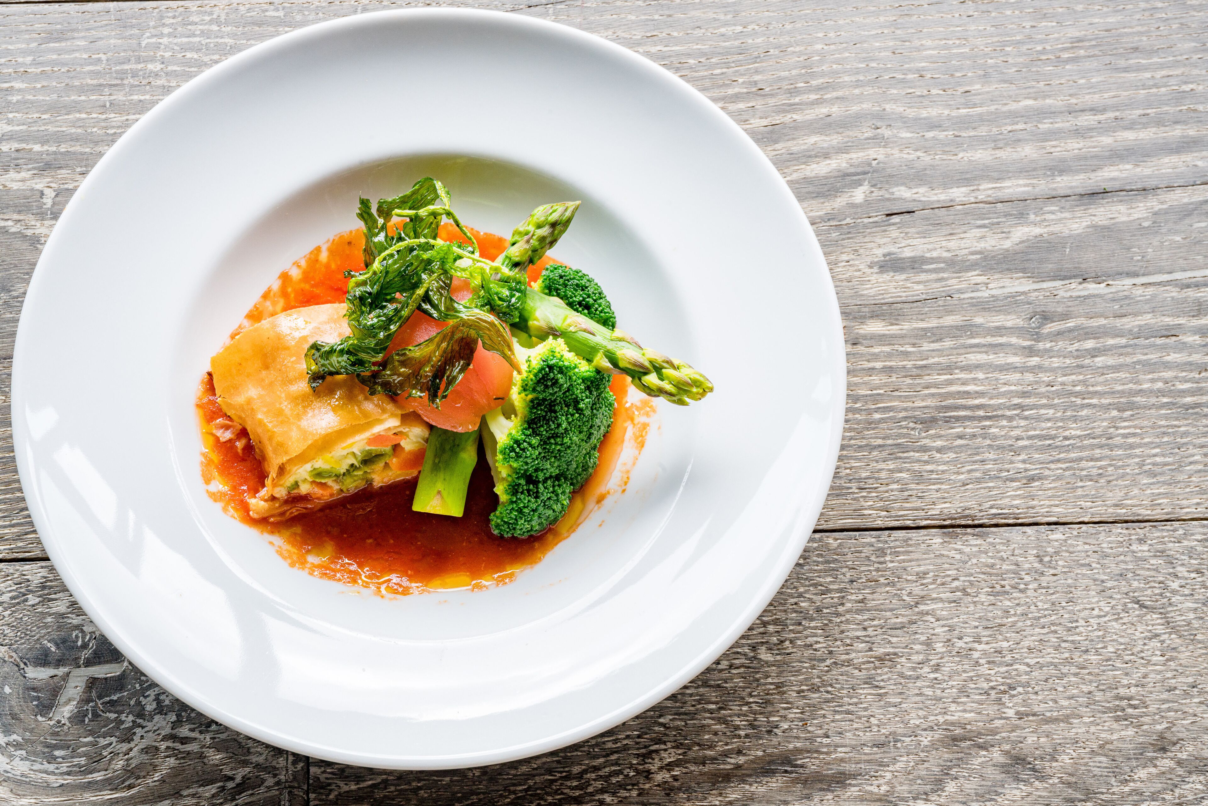 Une assiette blanche contenant un plat composé de légumes et d’une sauce à base de tomates, posée sur une table en bois.