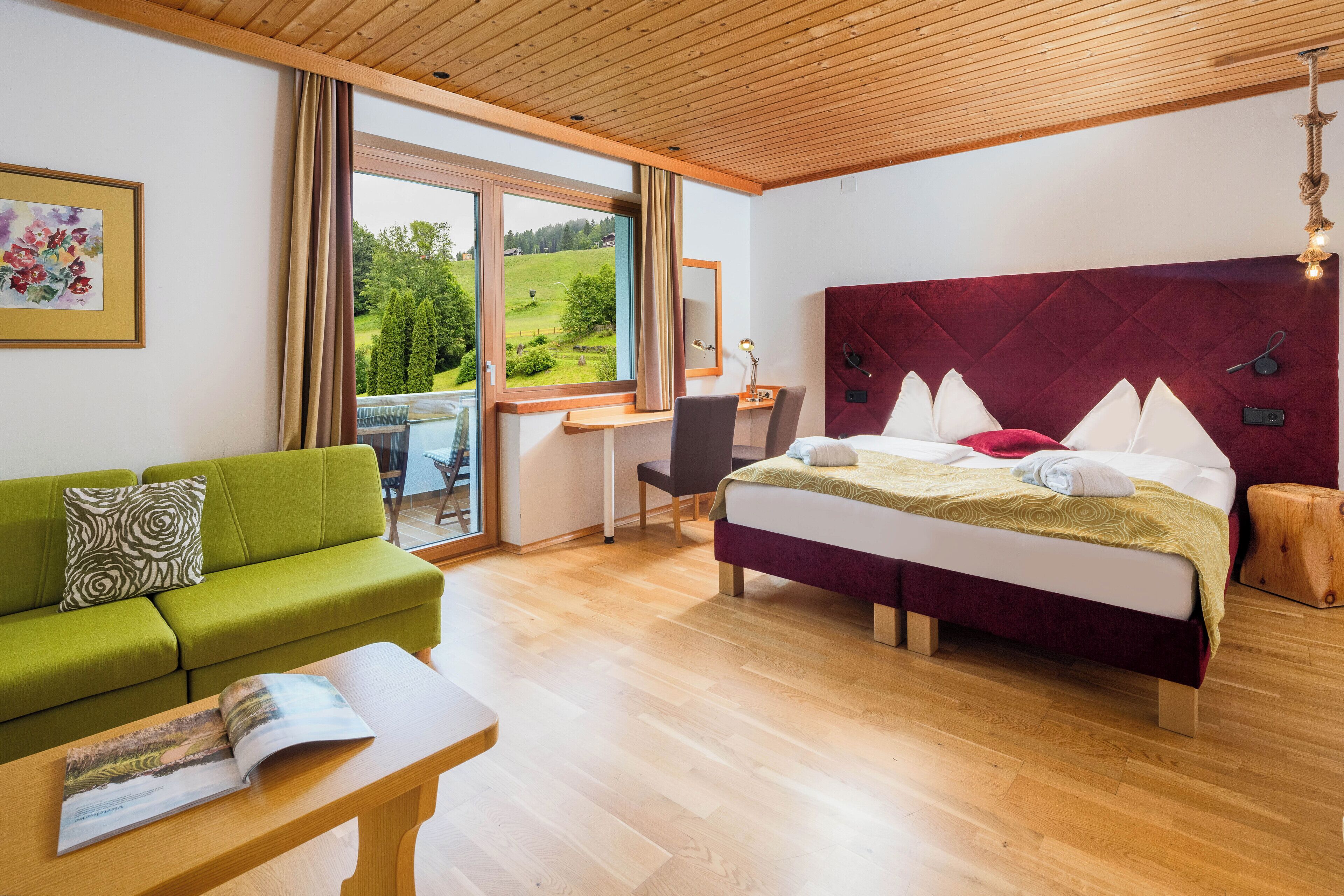 Habitación de hotel moderna con una cama grande, un sofá verde, un escritorio de madera y vistas al campo.