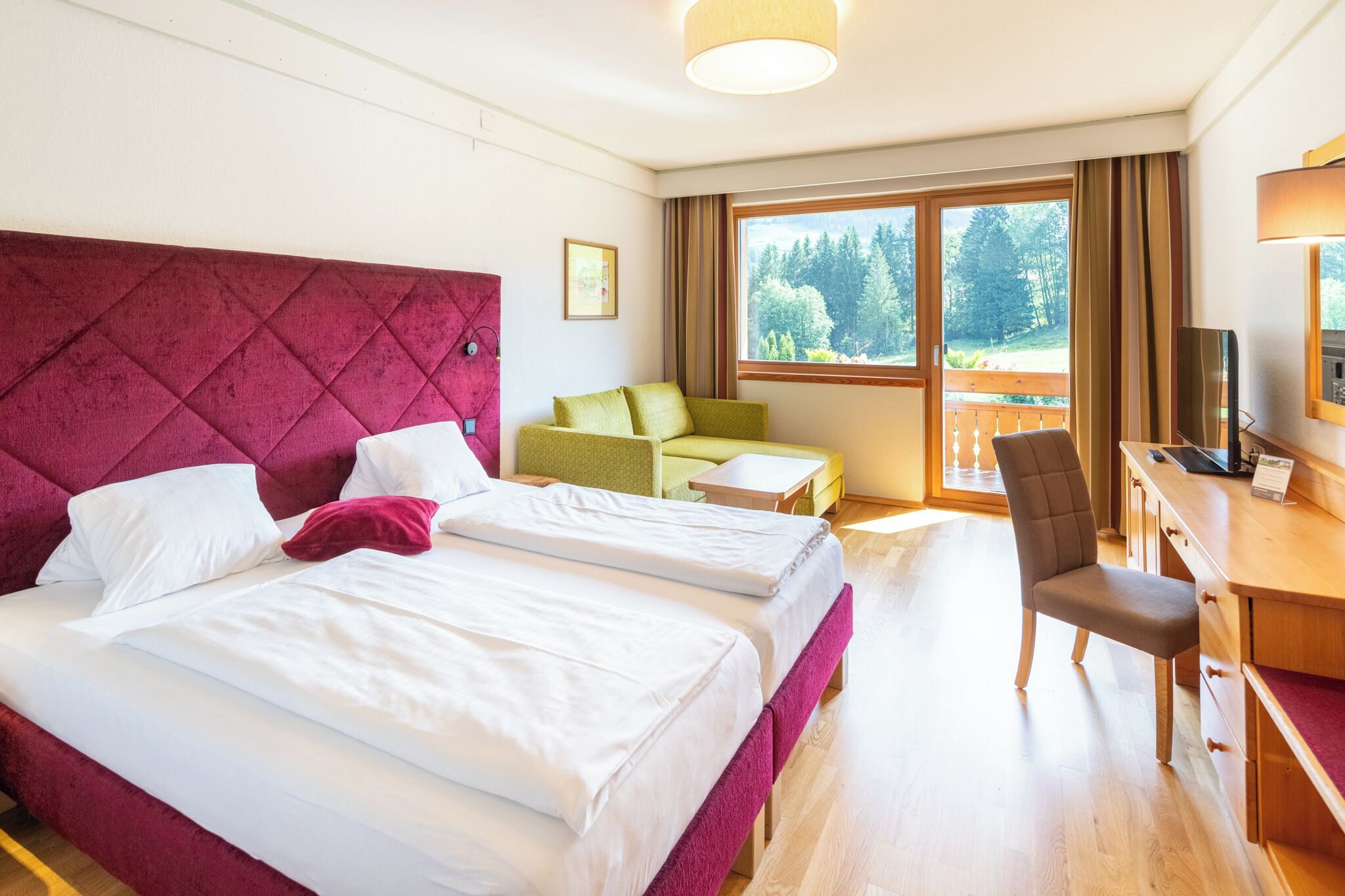 A hotel room with two beds, a desk, a chair, a TV, and a large window with a view of trees.