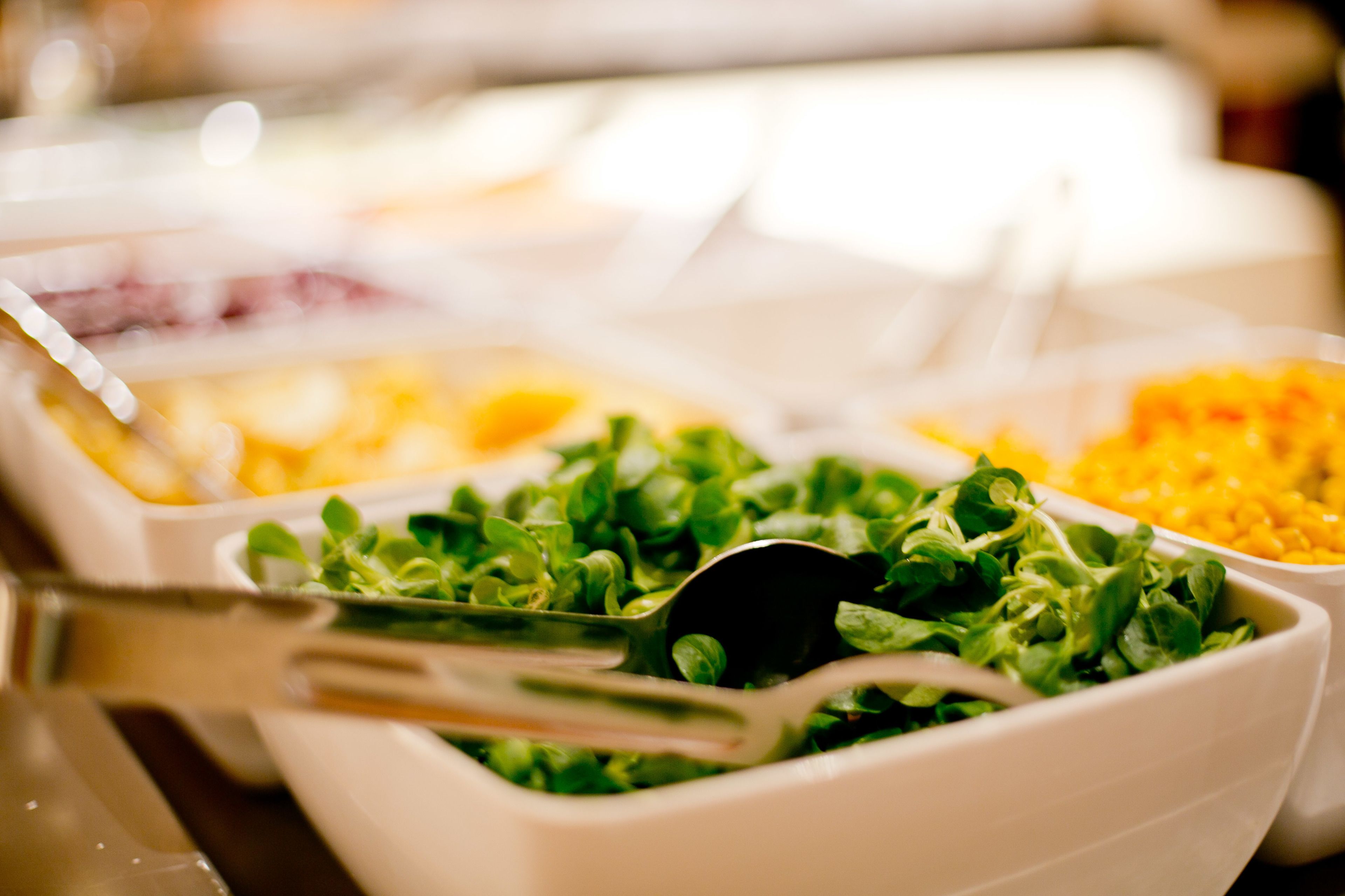 Un bufé con diversas opciones de comida, incluyendo un tazón de ensalada verde y un recipiente con maíz amarillo.