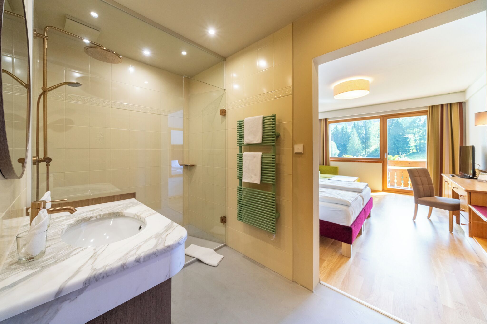 A modern bathroom with a large sink, shower, and a glass door leading to a bedroom with a bed and a desk.