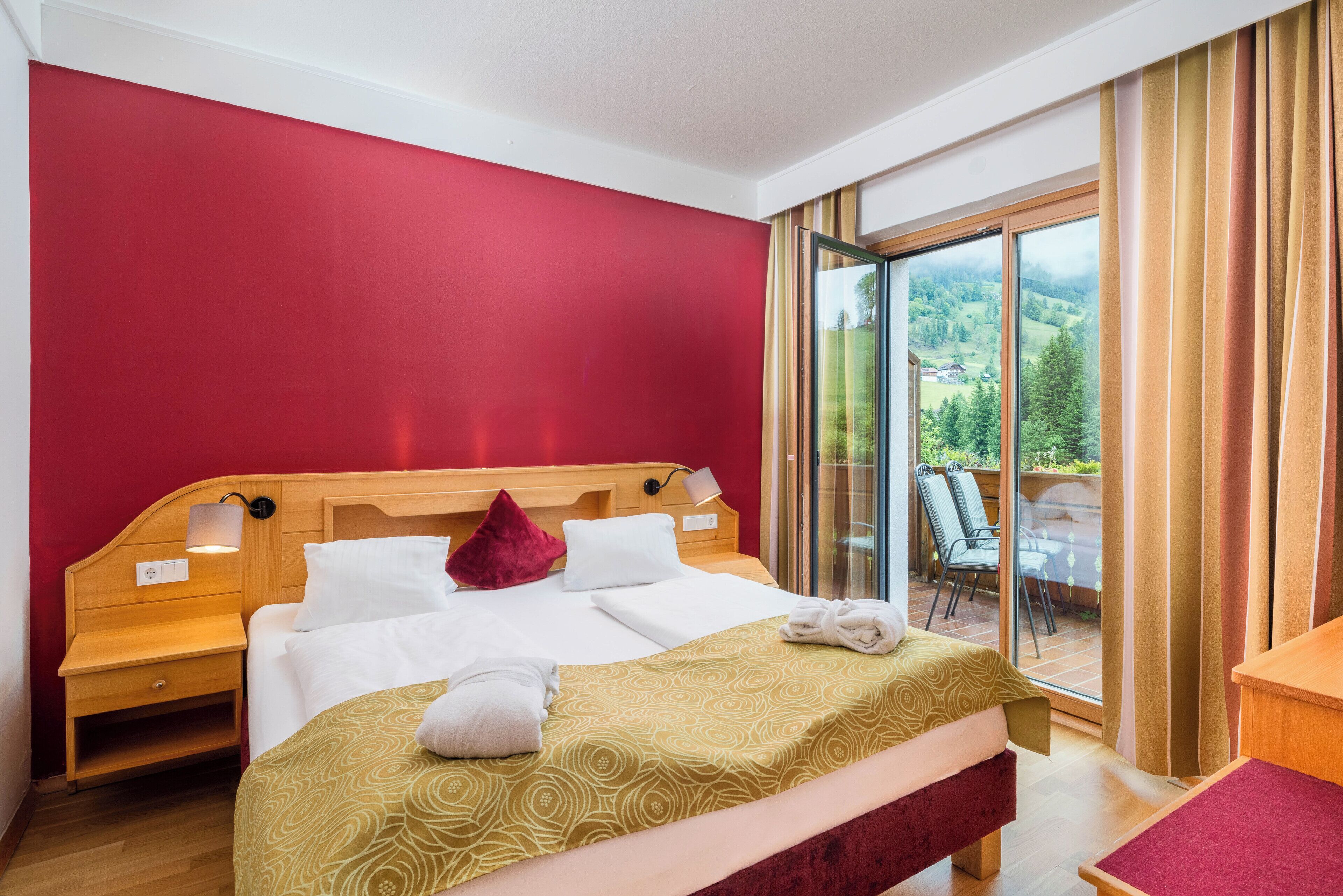 Une chambre d’hôtel avec un grand lit, une tête de lit en bois, un mur rouge et une porte-fenêtre coulissante donnant sur un espace extérieur aménagé avec des chaises.