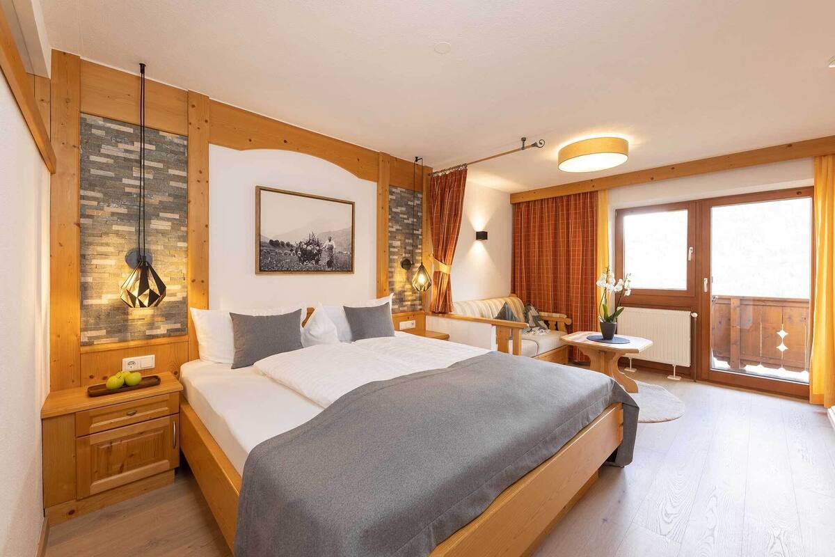 Un dormitorio amplio con una cama grande, cabecera de madera, mesita de noche con fruta, mesita auxiliar, lámpara de pared, un cuadro y vistas al exterior a través de una ventana.