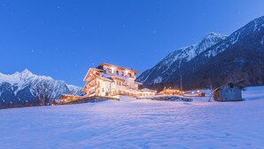 A snow-covered landscape with a large, illuminated building and a smaller structure, surrounded by mountains. - Aktiv Panoramahotel Daniel (Sautens)