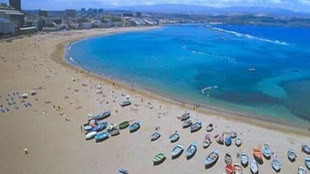 Una playa con numerosos barcos alineados en la arena, un mar azul cristalino y edificios al fondo.