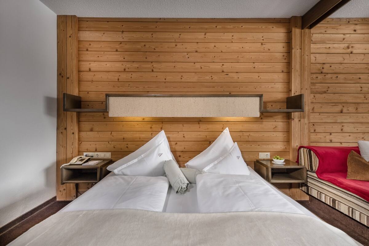 Une chambre d’hôtel avec une tête de lit en bois, un lit recouvert de draps blancs, un canapé rouge et une petite table sur laquelle se trouve un saladier rempli de fruits.