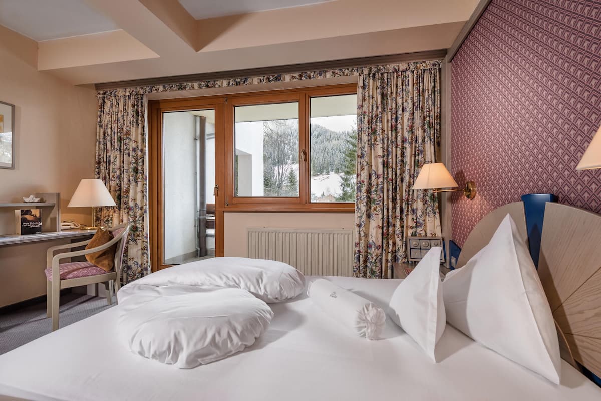 Une chambre d’hôtel avec un lit, deux lampes, un bureau, une chaise et une fenêtre avec des rideaux.