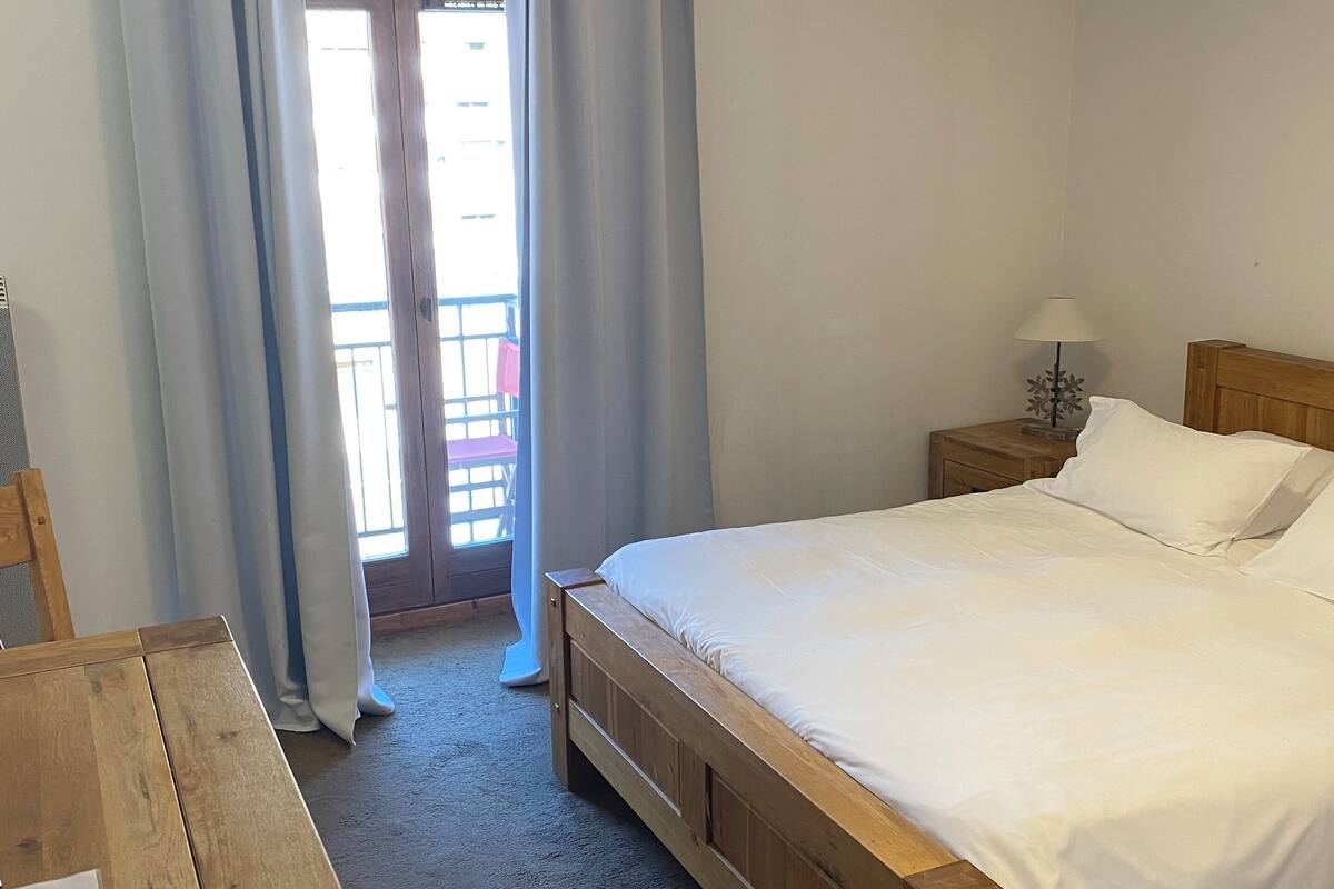 Une chambre à coucher avec un lit en bois, une table de chevet avec une lampe, un bureau en bois et une vue sur l’extérieur par une fenêtre ornée de rideaux.