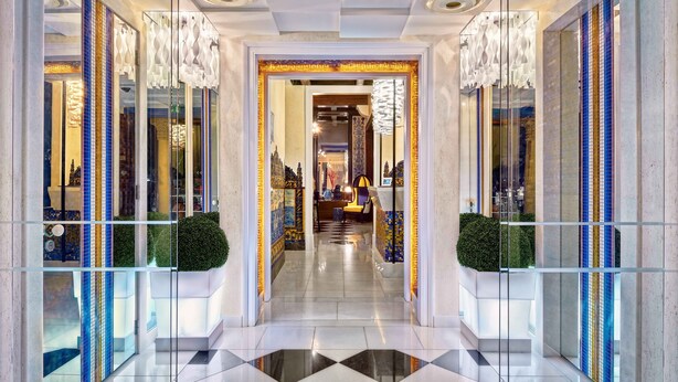 A modern hotel lobby with a glass partition, a tiled floor, and a central chandelier.