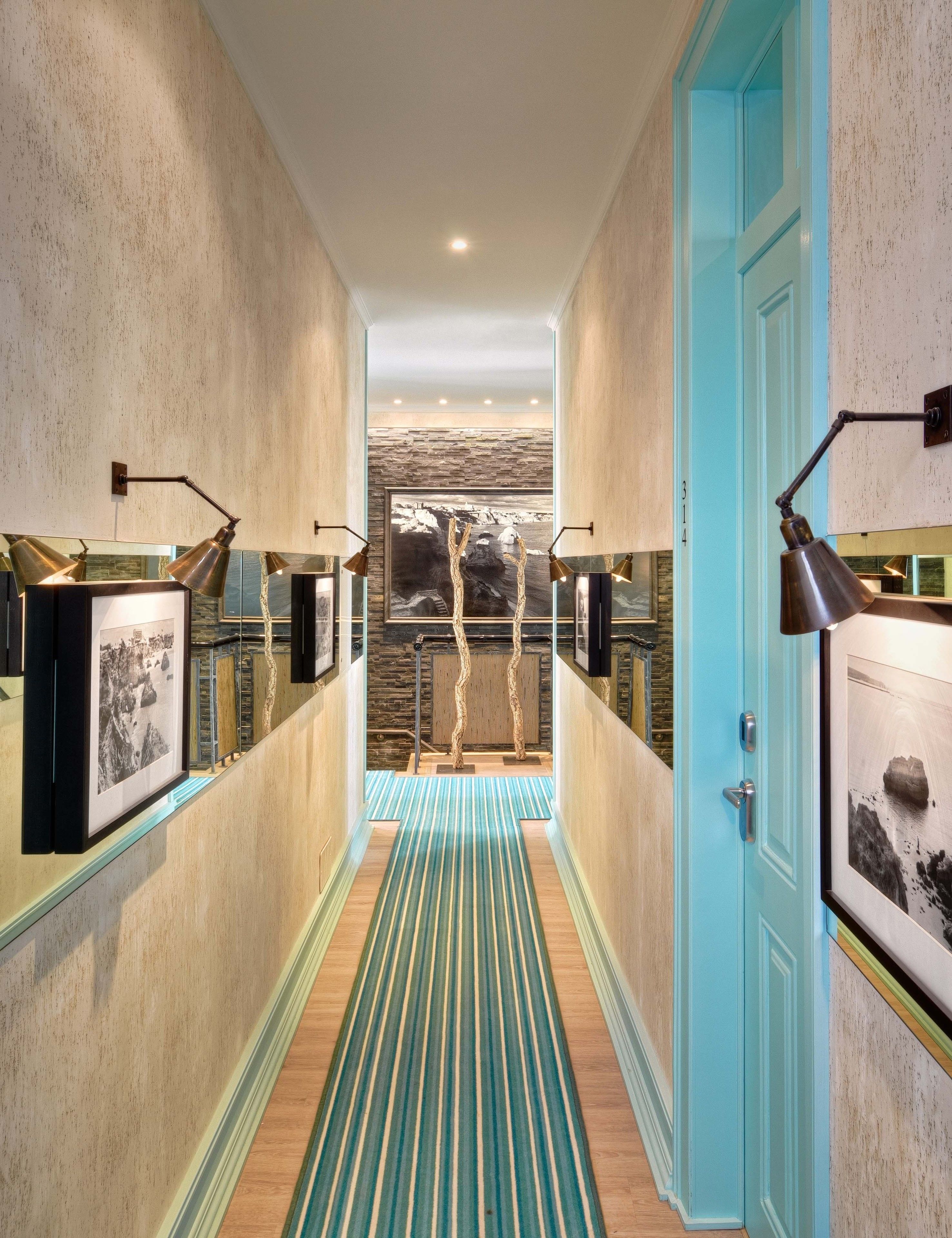 Un couloir avec un tapis à rayures, des tableaux encadrés accrochés aux murs et une porte bleue au fond.