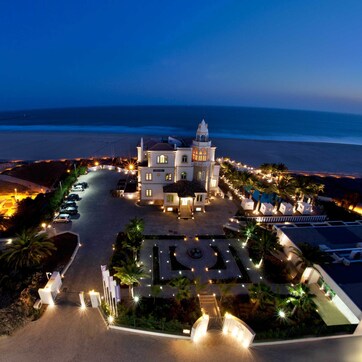 An aerial view of a coastal hotel with a central building, a beach, and a parking area.
