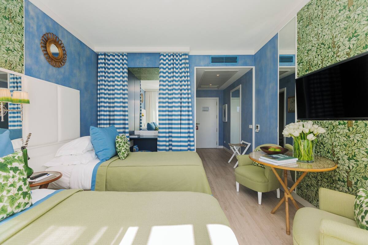 A hotel room with two beds, a small table with a vase of flowers, a green armchair, and a television on the wall.