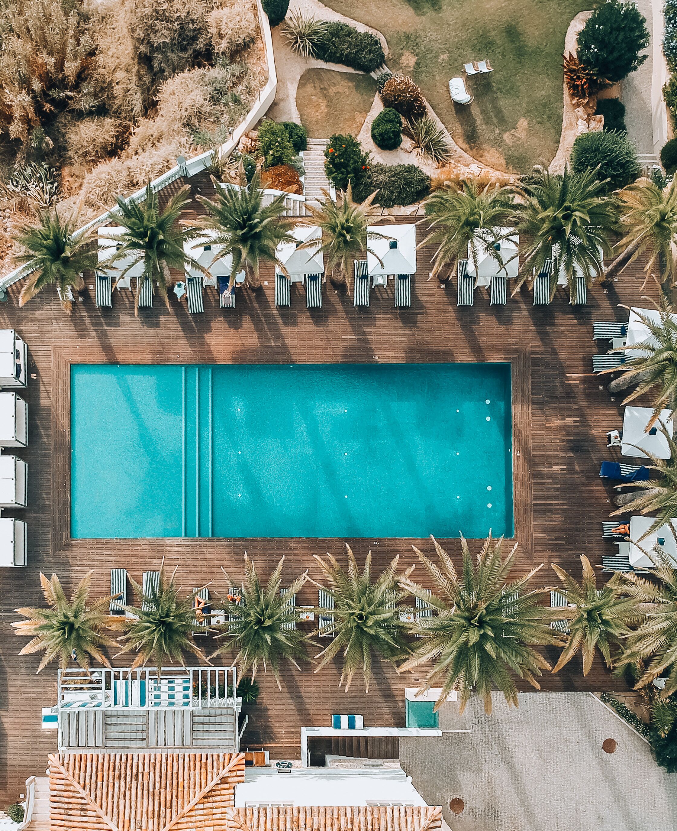 Vista aérea de um resort com uma piscina retangular grande, espreguiçadeiras e palmeiras.