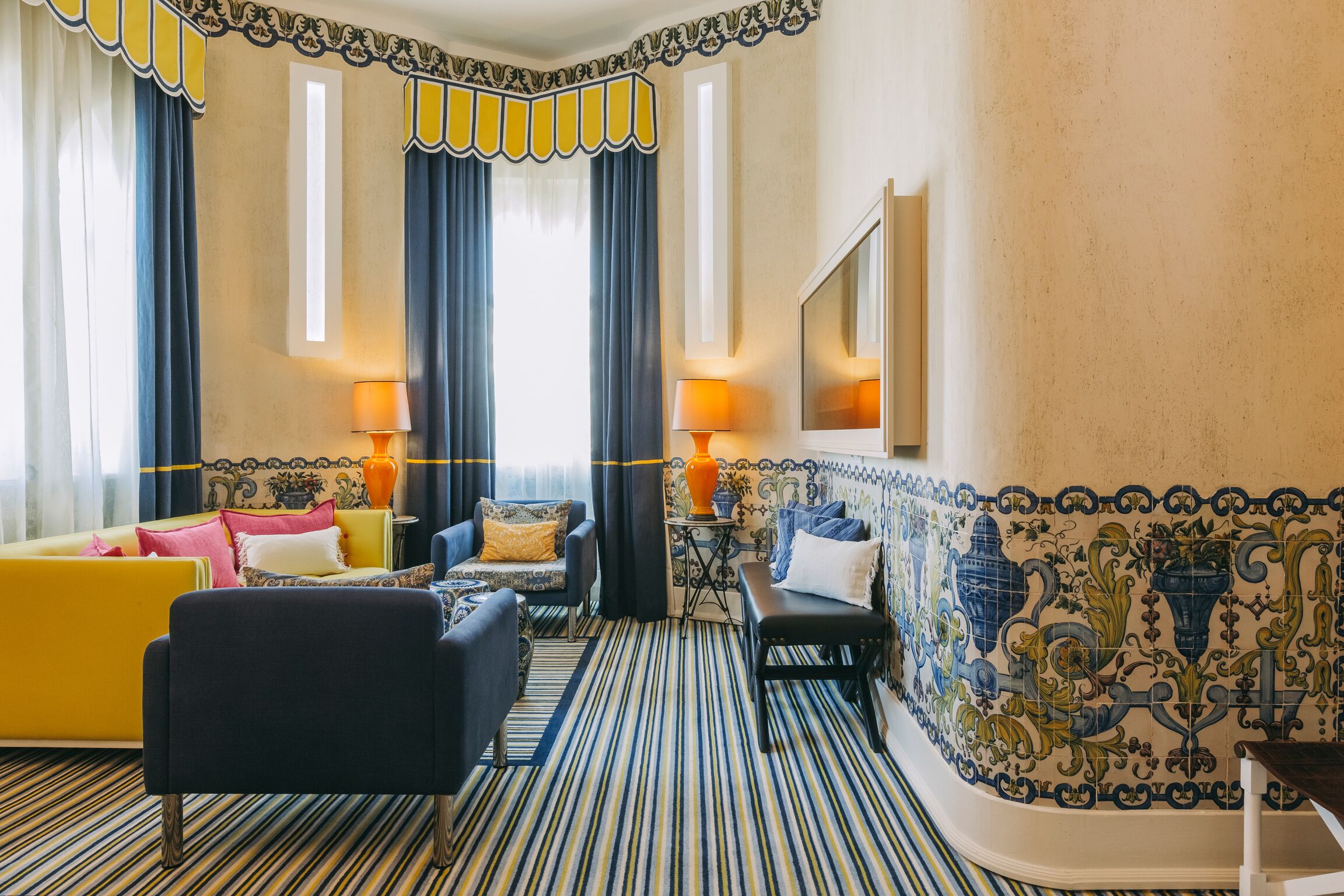 A hotel room with striped carpet, a yellow sofa, a blue armchair, a black bench, and a tiled wall with decorative patterns.