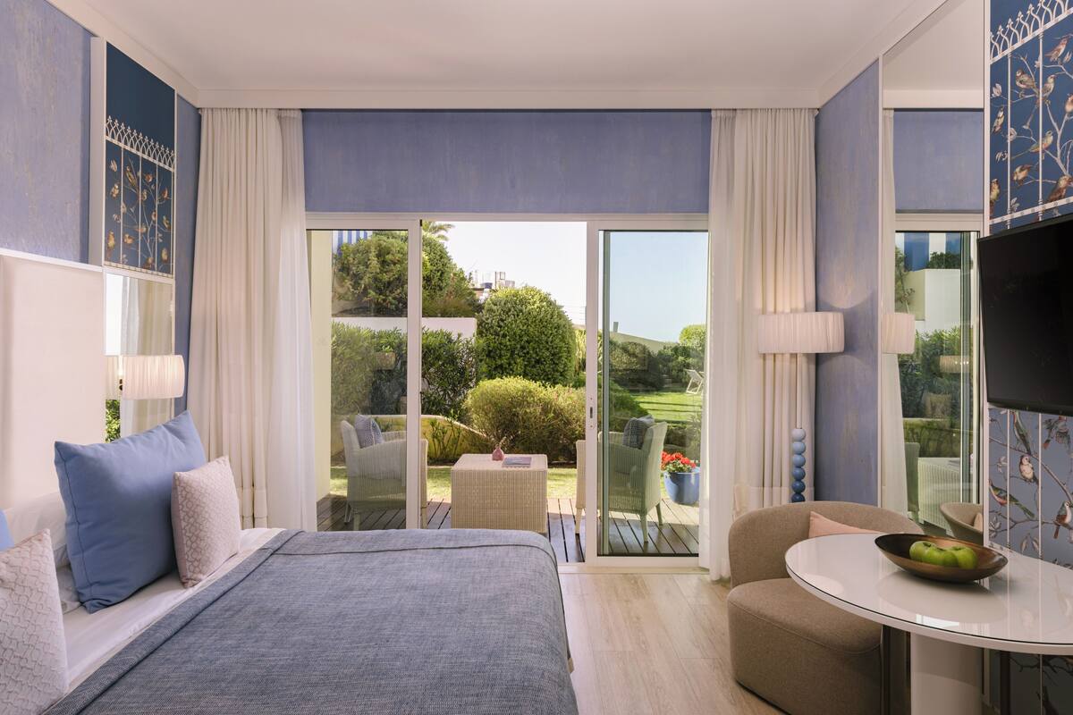 A modern hotel room with a large bed, a round dining table, a sofa, and a balcony with a view of the garden.