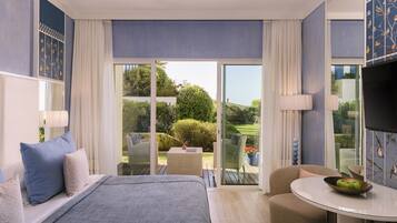 A modern hotel room with a large bed, a round dining table, a sofa, and a balcony with a view of the garden.