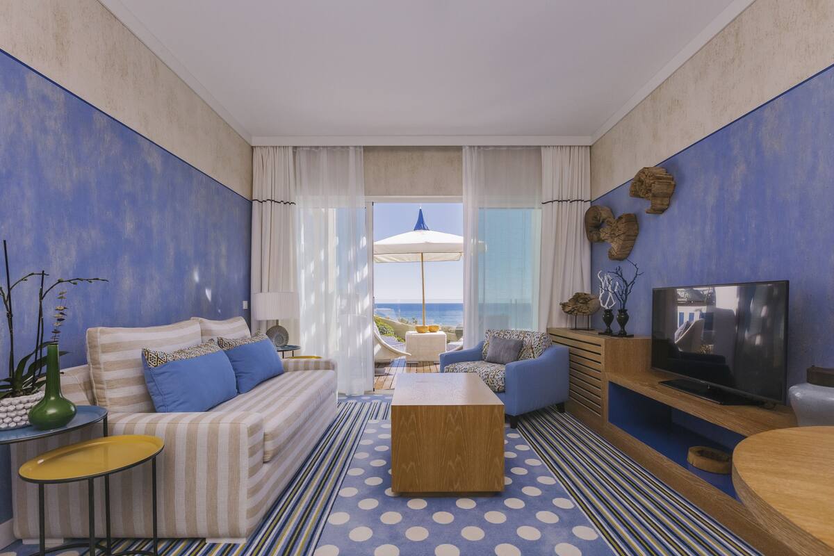 A modern living room with a sofa, coffee table, and TV, featuring a view of the ocean through large windows.
