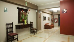 A hotel lobby with a polished tile floor, a wooden chair, a cushioned bench, and a mirror with a vase of flowers. - Hotel Kamana (Lima)