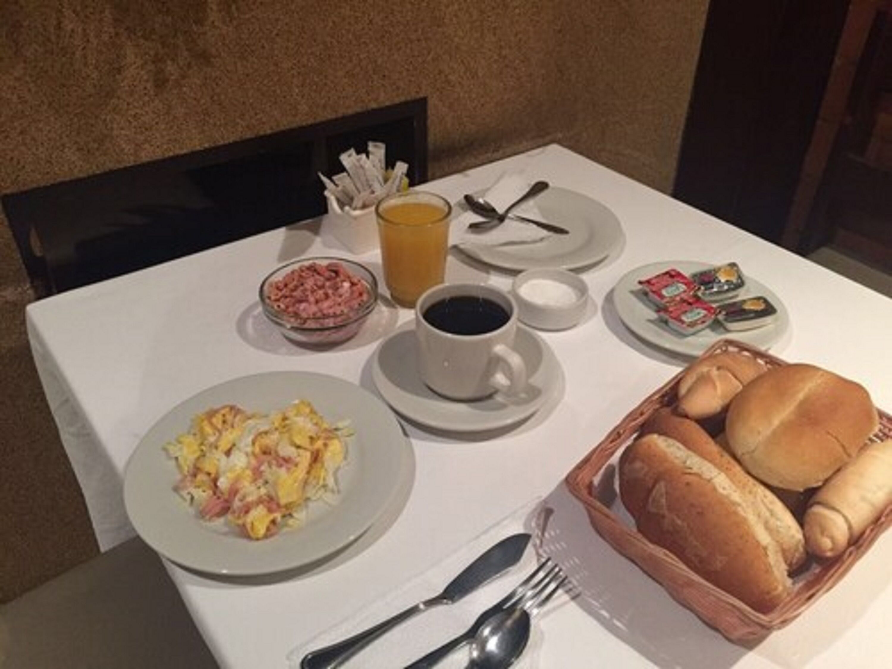 Una mesa de desayuno con platos de huevos revueltos, un bol de jamón, un vaso de zumo de naranja, una taza de café y una cesta de panecillos.