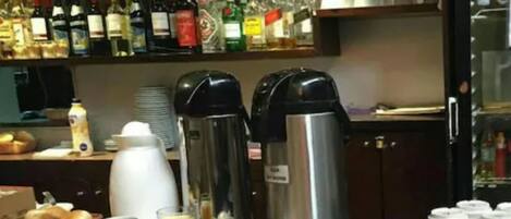 Mesa posta para o café da manhã, com máquinas de café, cesta de pão e diversas opções.