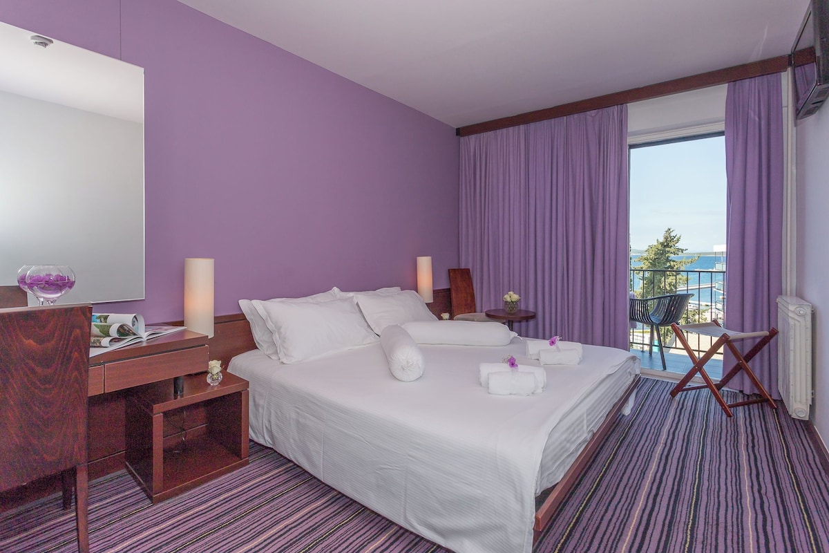 A hotel room with a large bed, a desk, a chair, and a view of the sea.
