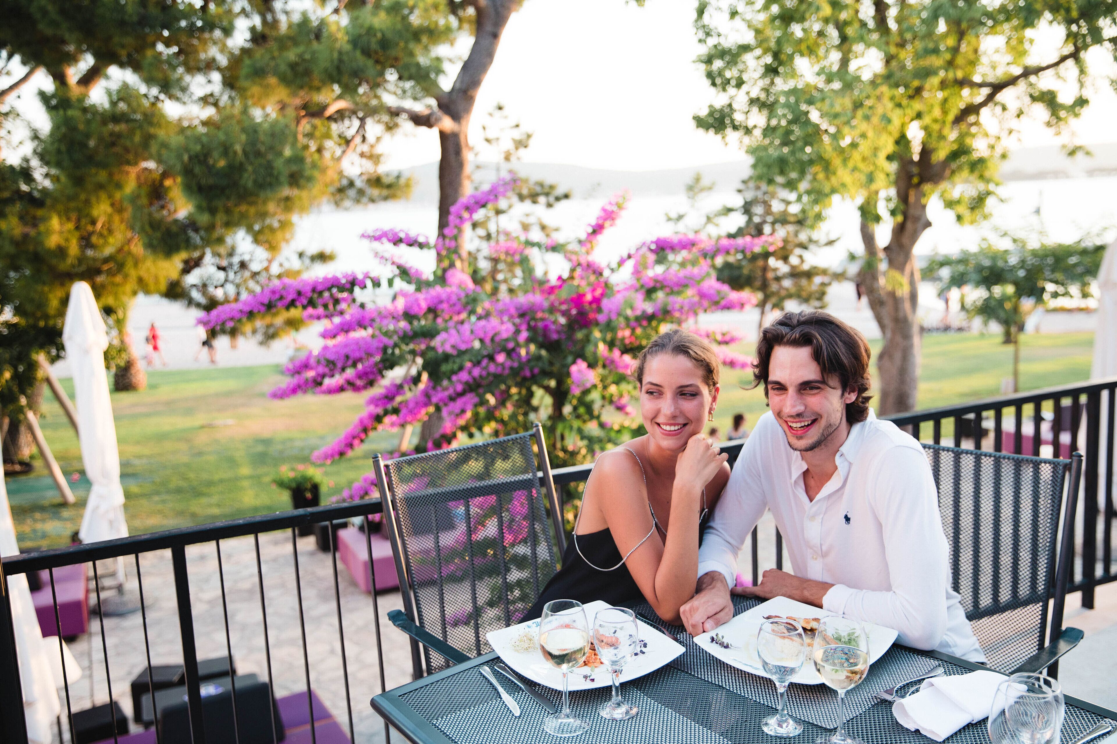 A couple dining outdoors with a table set for two, surrounded by lush greenery and purple flowers.