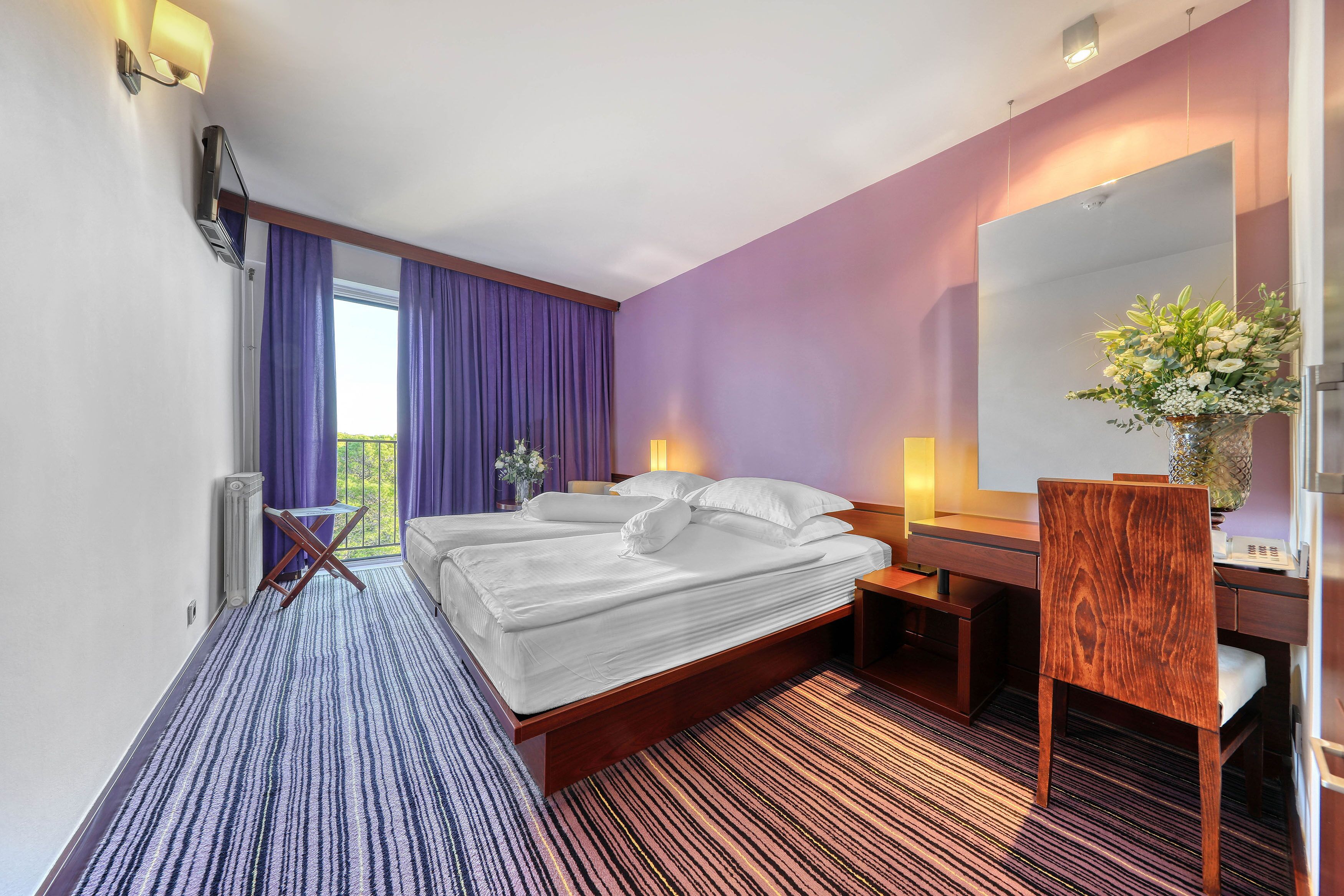 A hotel room with a large bed, a desk, a chair, a mirror, and a window with purple curtains.