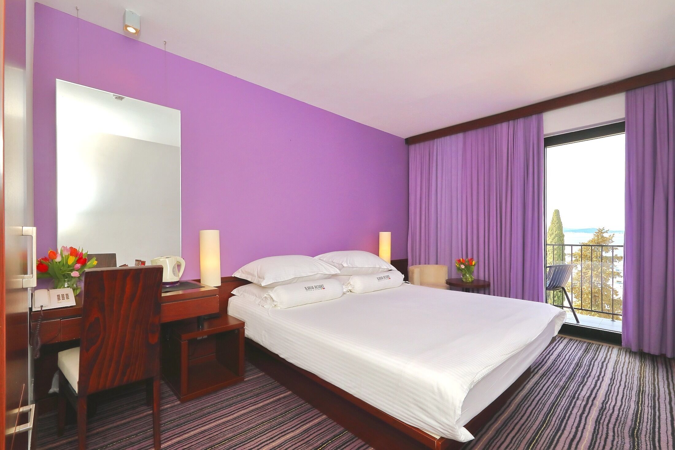 Habitación de hotel con una cama grande, un escritorio con teléfono, una silla y una mesita de noche con lámpara. Se ve una ventana con cortinas moradas y vistas al exterior.