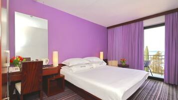 Habitación de hotel con una cama grande, un escritorio con teléfono, una silla y una mesita de noche con lámpara. Se ve una ventana con cortinas moradas y vistas al exterior.